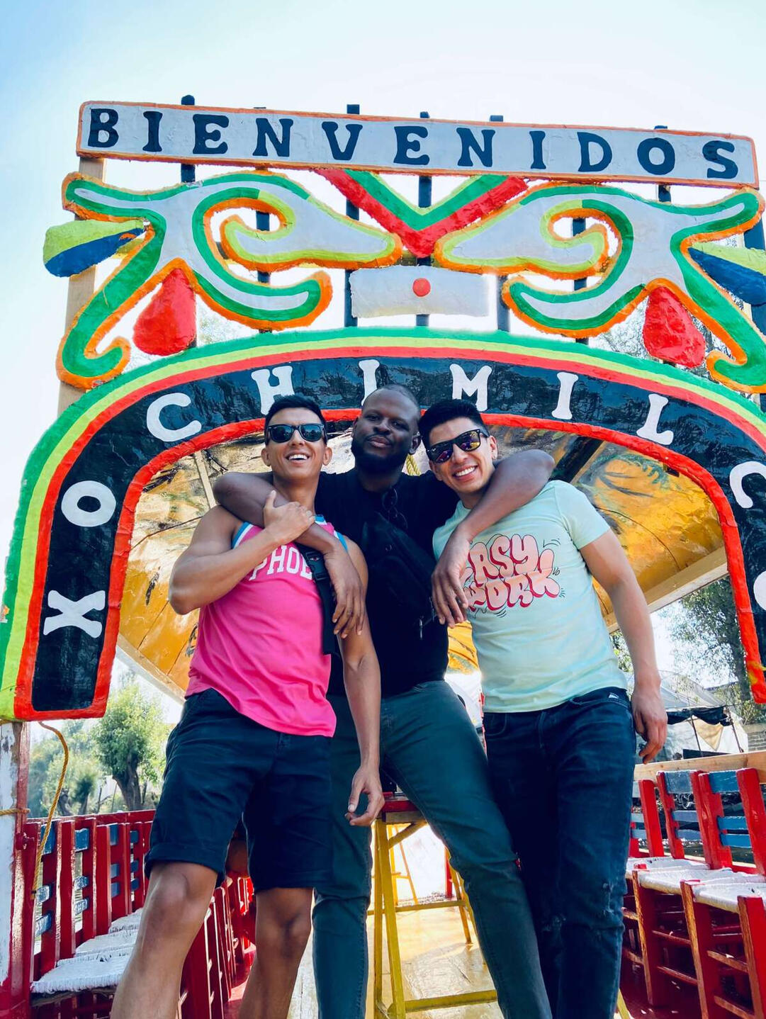 Erik and tourist posing under the colorful 'Bienvenidos a Xochimilco' arch of a traditional trajinera boat, in Mexico City.